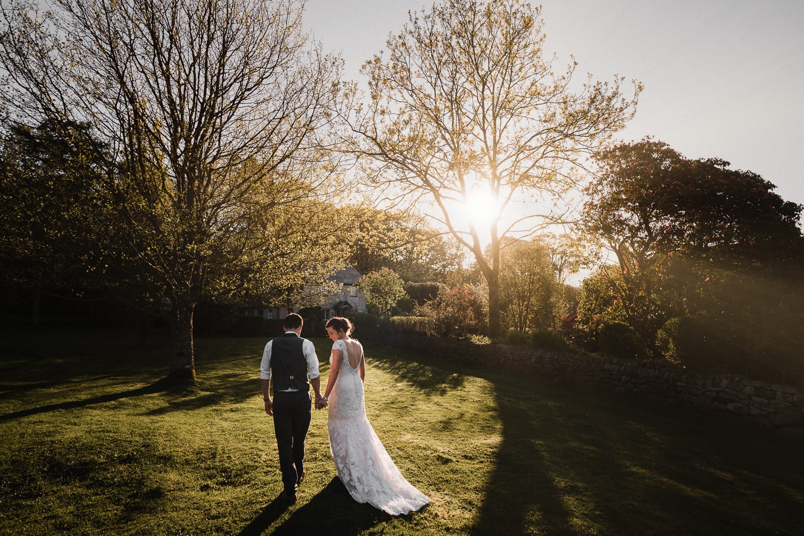 Bride and groom strolling hand in hand through the grounds of Cosawes Barton on their wedding day, while the sun sets through the trees